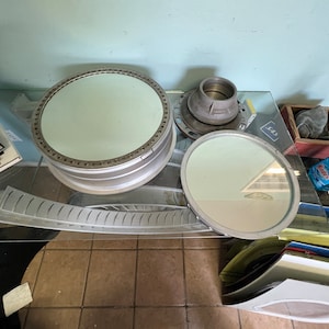 May include: Three round, silver-colored mirrors with reflective surfaces. The mirrors are on a glass table with a silver-colored metal object in the background.