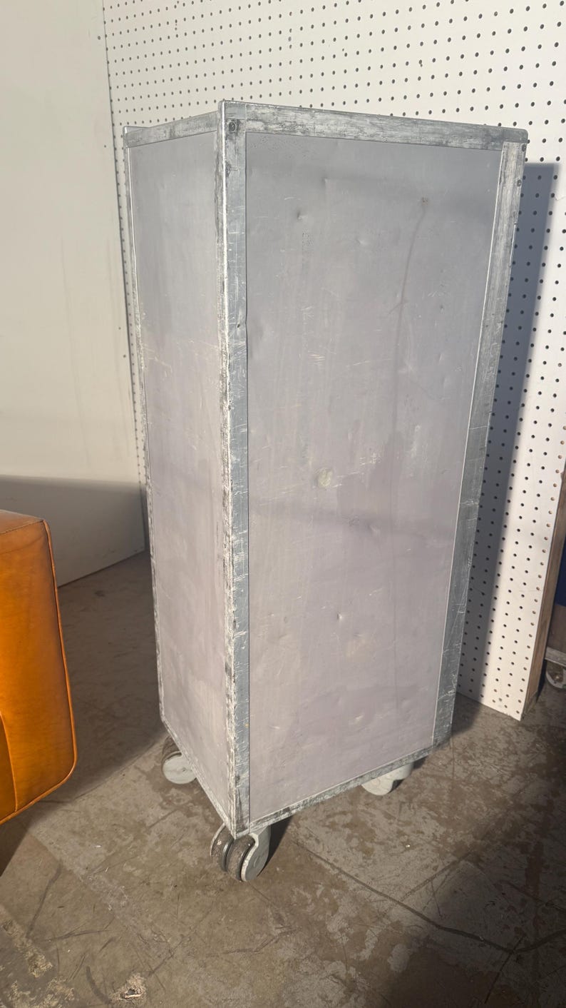 K&ouml;nnte beinhalten: Ein hoher, silberner Metallschrank mit vier R&auml;dern. Der Schrank hat eine rechteckige Form und eine glatte, leicht strukturierte Oberfl&auml;che. Der Schrank steht auf einem Betonboden.