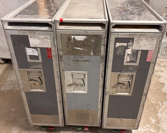 May include: Three industrial-style metal storage cabinets with a weathered appearance. Each cabinet has a rectangular shape, a handle, and a door. The cabinets are gray and silver, with some stickers and wear visible. The cabinets are on wheels.