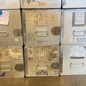 May include: A stack of silver metal storage containers with handles and latches. Some containers have labels and stickers from airlines such as Ryanair, LATAM, and Saudia. The containers are likely used for transporting goods.
