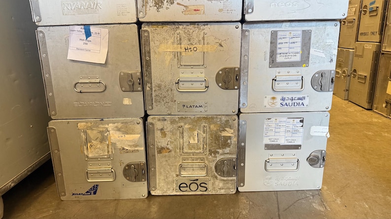 May include: A stack of silver metal storage containers with handles and latches. Some containers have labels and stickers from airlines such as Ryanair, LATAM, and Saudia. The containers are likely used for transporting goods.