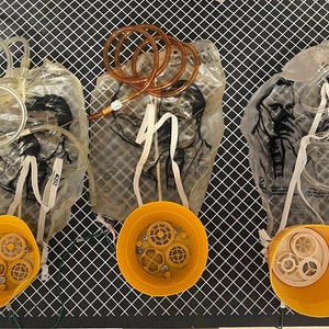 May include: Three yellow oxygen masks with clear plastic bags and tubing. The bags have a graphic of a person's face. The masks are on a black and white grid surface.