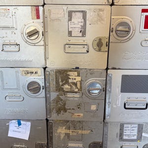 May include: A stack of silver metal storage containers with various labels, handles, and circular locking mechanisms. Some containers have red or gray accents. The containers are rectangular and appear to be industrial or commercial.