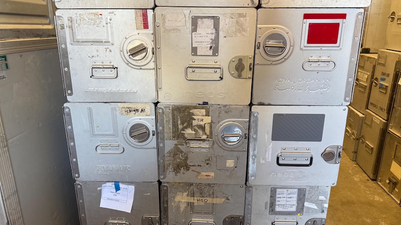 May include: A stack of silver metal storage containers with various labels, handles, and circular locking mechanisms. Some containers have red or gray accents. The containers are rectangular and appear to be industrial or commercial.