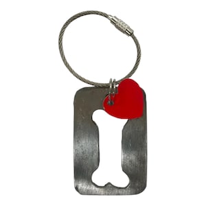 May include: A metal dog bone-shaped keyring with a red heart charm. The keyring has a silver-coloured wire loop for attaching to keys. The bone shape is cut out of the metal.