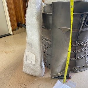 May include: A close-up of a large, gray metal part with a yellow measuring tape. The part is about 12 inches long and has a series of holes along the side. The part is sitting on a concrete floor.