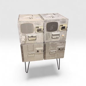May include: A storage unit constructed from four stacked silver-colored metal airline galley carts. The unit features industrial latches, handles, and ventilation grilles on the front panels, supported by four thin black metal hairpin legs.