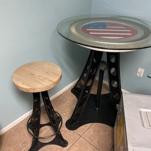 Puede incluir: Un taburete de bar de metal negro con un asiento de madera y una mesa redonda de vidrio con una base de metal negro. La tapa de la mesa presenta un diseño circular con una bandera estadounidense roja, blanca y azul.