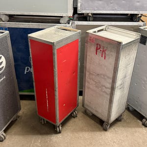 May include: A collection of used airline food service carts. The carts are rectangular with wheels, and made of metal. One cart is red, and another has a logo and the word "NEWORK" on the side. Some carts have the letters "P-K" written on them.