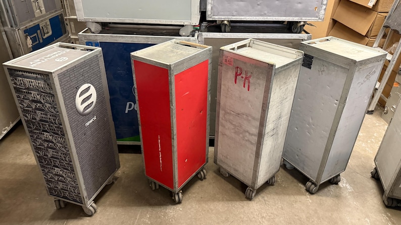 May include: A collection of used airline food service carts. The carts are rectangular with wheels, and made of metal. One cart is red, and another has a logo and the word "NEWORK" on the side. Some carts have the letters "P-K" written on them.