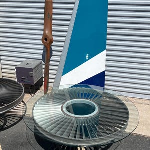 May include: A glass-top coffee table with a unique design featuring a large, circular metal base that resembles an airplane engine. The base is painted in shades of gray and silver, and the glass top is clear. The table is surrounded by a white line on a black asphalt surface.