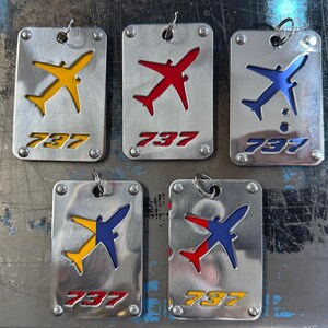 May include: Five silver keychains featuring a 737 airplane silhouette in various colour combinations. The airplanes are yellow and red, red and blue, blue and yellow, and blue and red. Each keychain has the number 737 printed on it.