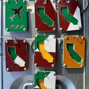 May include: Six metal keychains with different designs. One keychain is green with a white airplane and the text '737'. The other five keychains are red, white, green, and yellow with a cut-out of the state of California.