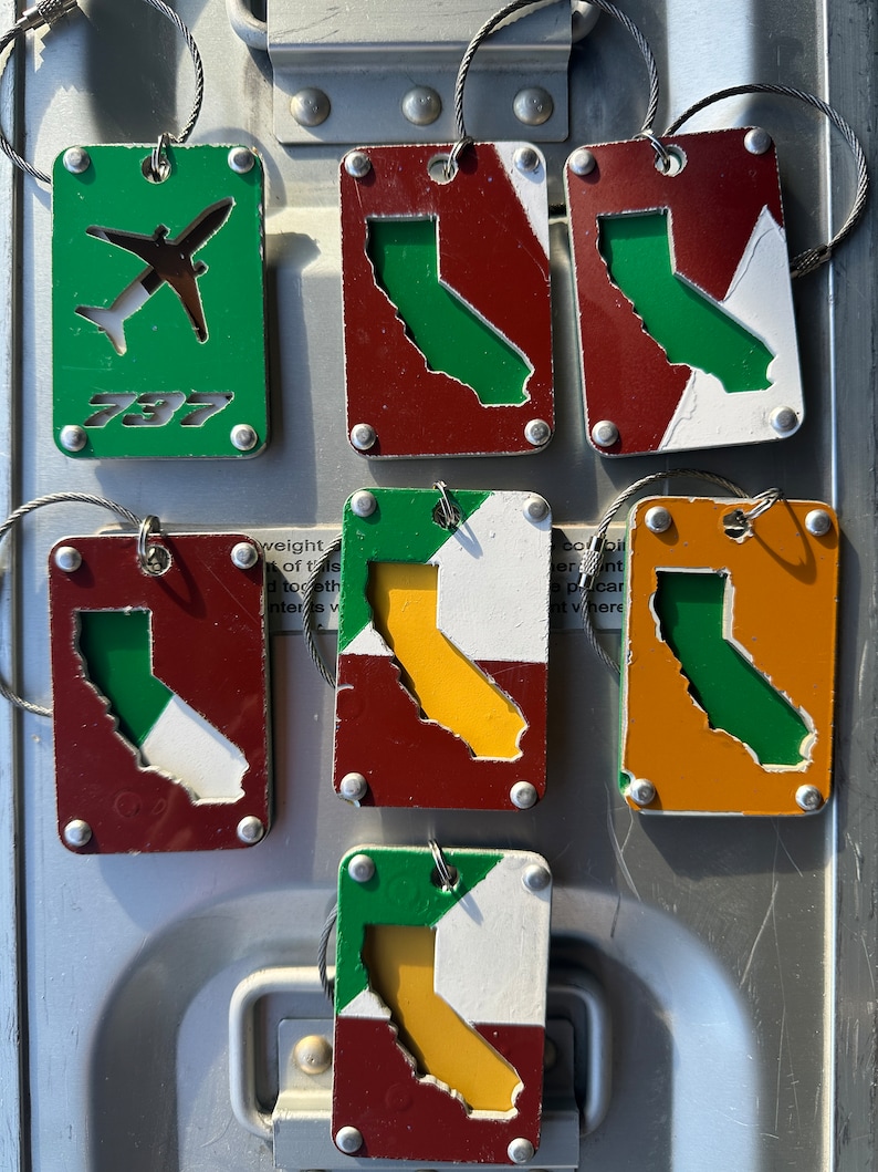 May include: Six metal keychains with different designs. One keychain is green with a white airplane and the text '737'. The other five keychains are red, white, green, and yellow with a cut-out of the state of California.