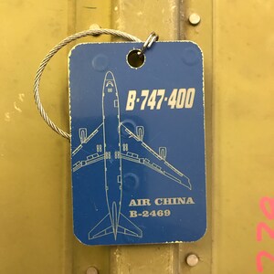 K&ouml;nnte beinhalten: Blaues Gep&auml;ckanh&auml;nger mit einer wei&szlig;en Umrandung eines Boeing 747-400 Flugzeugs. Der Anh&auml;nger hat den Text "B-747-400" und "AIR CHINA B-2469".