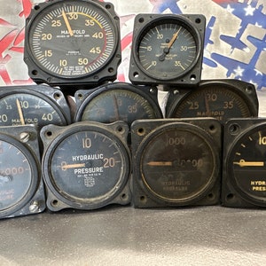 May include: A collection of nine vintage aircraft gauges, including manifold pressure gauges and hydraulic pressure gauges. The gauges are black with white markings and needles. Some gauges have a yellow background.