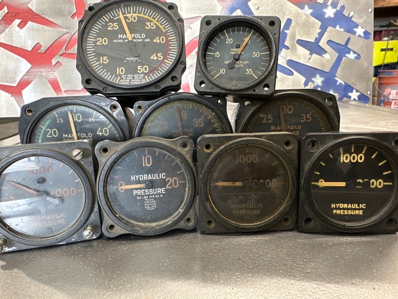 May include: A collection of nine vintage aircraft gauges, including manifold pressure gauges and hydraulic pressure gauges. The gauges are black with white markings and needles. Some gauges have a yellow background.