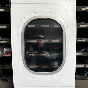 May include: A white airplane door with a large oval window. The window is reflecting a person standing in front of a wall of shelves with various labels. The labels include "Crosses", "Cross Large", "Cross Med", "Boeing Centers", "Angel Wings", "Texas", "Nevada", "California", "Circle 8737", "DC10 KF", "DGE Lug To", "Med Hearts", "W/F Narrow Rect", "FL".