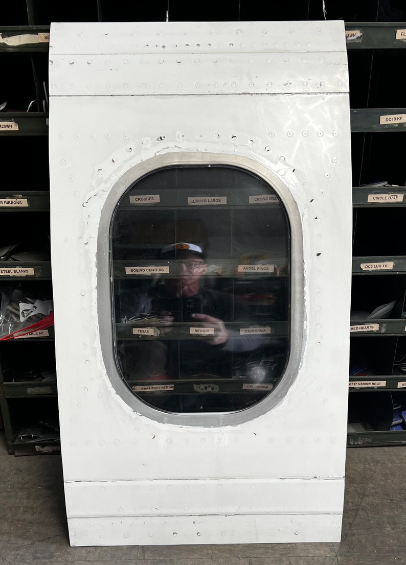 May include: A white airplane door with a large oval window. The window is reflecting a person standing in front of a wall of shelves with various labels. The labels include "Crosses", "Cross Large", "Cross Med", "Boeing Centers", "Angel Wings", "Texas", "Nevada", "California", "Circle 8737", "DC10 KF", "DGE Lug To", "Med Hearts", "W/F Narrow Rect", "FL".