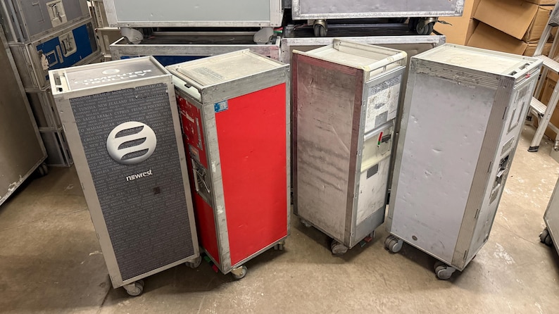 May include: Four industrial metal carts with wheels. One cart has a gray textured front with a white logo and the word "newest". Another cart is red. The other two carts are silver. They appear to be used for storage or transport.