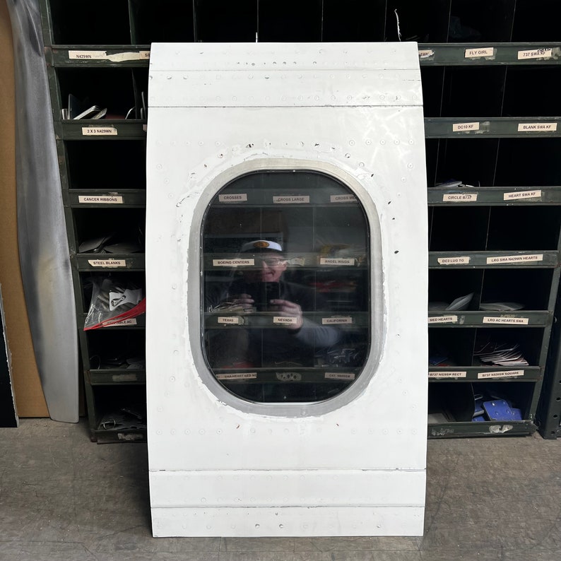 May include: A white airplane window with a round frame. The window is set into a white panel. A person is reflected in the window.