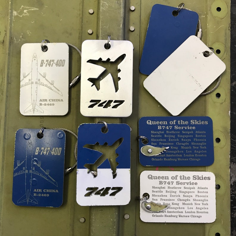 K&ouml;nnte beinhalten: F&uuml;nf Gep&auml;ckanh&auml;nger in blau-wei&szlig;er Farbgebung. Die Anh&auml;nger zeigen eine Silhouette eines Boeing 747 Flugzeugs und den Text "747" und "Queen of the Skies B747 Service". Die Anh&auml;nger enthalten auch den Text "Air China B-2469" und eine Liste der von der Fluggesellschaft bedienten St&auml;dte.