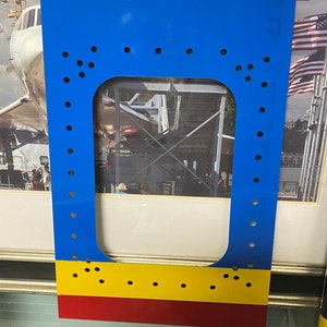 May include: A blue metal frame with a yellow and red stripe at the bottom. The frame has a rectangular cutout in the center and is decorated with a pattern of small holes.