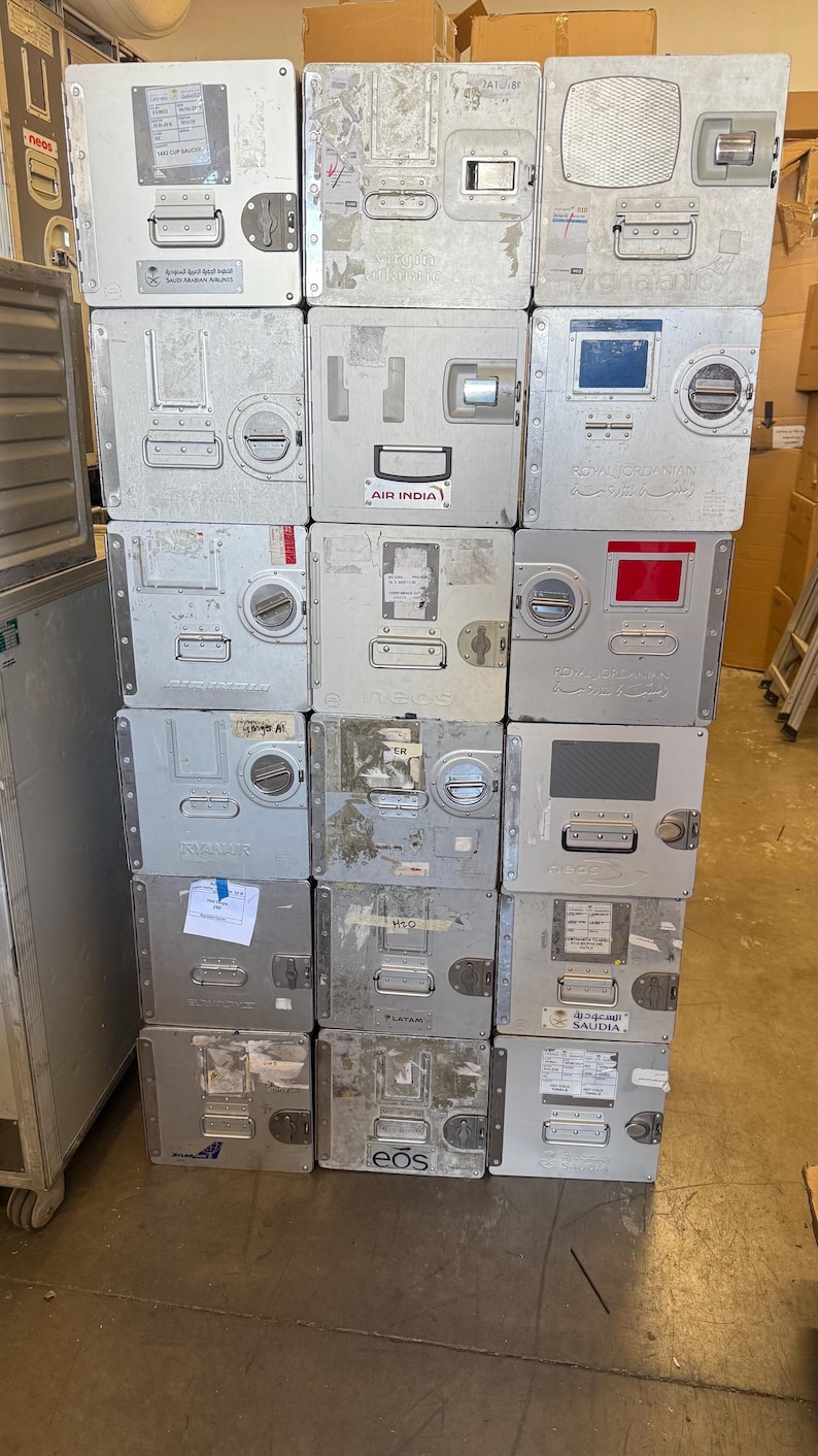 May include: A stack of used, silver-colored airline food service containers. Each box has a door, handle, and various labels and markings. Some boxes have the logos of airlines like Air India and Saudia.