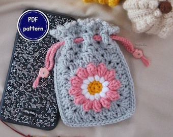 May include: A crocheted gray drawstring bag with a pink and white flower applique. The bag has a pink drawstring and a small pink flower accent. A phone case with a sparkly design is next to the bag. The text "PDF pattern" is in a circle.