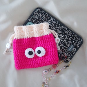 May include: A handmade, cerise crochet drawstring pouch with large white and black googly eyes. The top is cream-coloured. A bedazzled phone case and beaded lanyard are in the background.