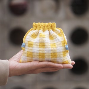Puede incluir: Pequeño bolso de ganchillo hecho a mano con cordón, con un patrón de cuadros amarillos y blancos. La bolsa tiene una parte superior amarilla y se cierra con un cordón amarillo y cuentas azul claro. La bolsa es lo suficientemente pequeña como para caber en la palma de una mano.
