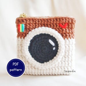 Könnte beinhalten: Gehäkelte Instagram-Kamera-Münzbörse in Braun, Weiß und Schwarz. Die Börse hat einen Reißverschluss und einen regenbogenfarbenen Streifen an der Seite. Der Text "PDF pattern" befindet sich auf einem blauen Kreis.