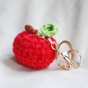 May include: A red crocheted apple keychain with a green leaf and a gold metal key ring.