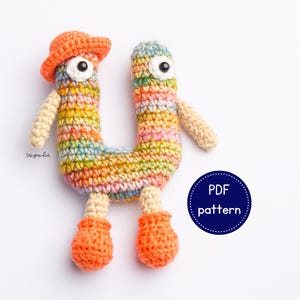 Puede incluir: Un juguete amigurumi de crochet con forma de la letra U. El juguete lleva un sombrero naranja brillante y tiene ojos grandes y blancos. El juguete está hecho con un arcoíris de colores, incluyendo rosa, azul, verde y amarillo. El juguete está de pie sobre una superficie blanca. Patrón PDF.