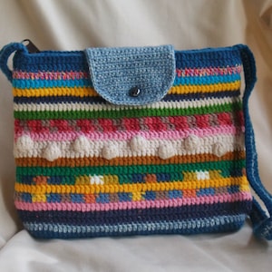 May include: A handmade crochet crossbody bag with a blue base and strap. The bag features a colorful striped pattern with various colors, including pink, yellow, green, and white. The flap is light blue and secured with a button.
