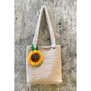 May include: A beige crocheted tote bag with a crocheted sunflower embellishment. The sunflower is yellow with a brown center and green leaves.