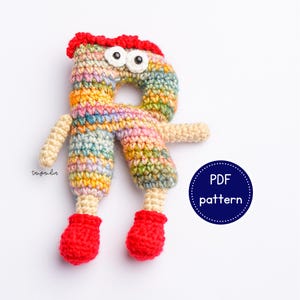 May include: A crocheted amigurumi toy shaped like the letter R. The toy is colourful with a rainbow pattern and has googly eyes, red shoes, and a red hairpiece. The text "PDF pattern" is visible in a blue circle.