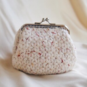 May include: A cream-colored, crocheted coin purse with a silver-toned clasp. The purse features a textured, star-like pattern with flecks of red and yellow. The clasp is a classic kiss-lock style.