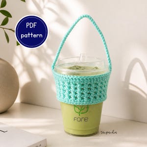 Peut inclure: Un gobelet en plastique transparent rempli d'une boisson verte, avec une housse et une poignée crochetées bleu clair. Le gobelet a un couvercle transparent et le mot "fore" est imprimé sur le gobelet. Un cercle bleu avec "PDF pattern" est en arrière-plan.