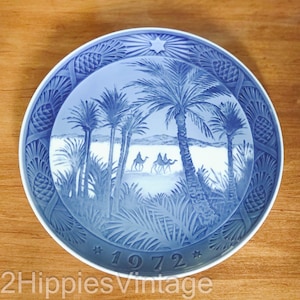 May include: A round porcelain decorative plate featuring a blue and white relief design of palm trees and camels in a desert landscape. The border has a textured pinecone pattern, and the bottom edge displays the year 1972 with two small stars.