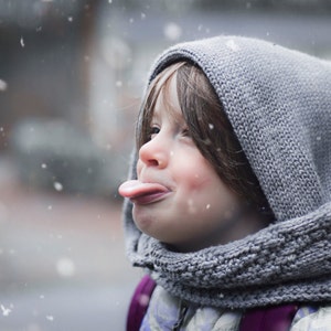 Puede incluir: Un ni&ntilde;o peque&ntilde;o con una bufanda con capucha gris y un su&eacute;ter saca la lengua mientras est&aacute; de pie en la nieve.