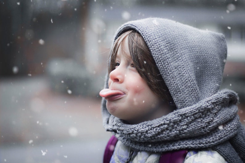 Puede incluir: Un ni&ntilde;o peque&ntilde;o con una bufanda con capucha gris y un su&eacute;ter saca la lengua mientras est&aacute; de pie en la nieve.