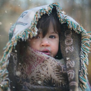Puede incluir: Un ni&ntilde;o peque&ntilde;o con una manta colorida con borde de flecos, mirando hacia arriba con una expresi&oacute;n seria. El ni&ntilde;o est&aacute; de pie en un entorno exterior nevado.