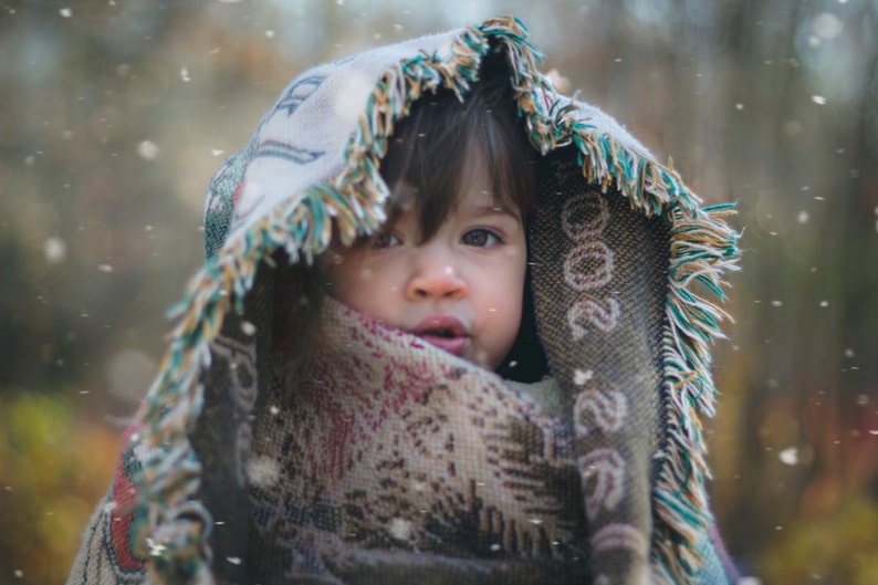 Puede incluir: Un ni&ntilde;o peque&ntilde;o con una manta colorida con borde de flecos, mirando hacia arriba con una expresi&oacute;n seria. El ni&ntilde;o est&aacute; de pie en un entorno exterior nevado.