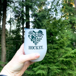 May include: White tumbler with a black graphic that says "You had me at hockey." The graphic includes a hockey stick, puck, and goalie mask.