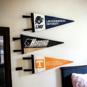 May include: Three triangular felt pennants displayed horizontally. The top pennant is blue and white with the Lincoln Memorial University logo. The middle is black and gold with the Harding University logo. The bottom is orange and white with the University of Tennessee logo.