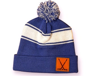 May include: A blue knitted beanie hat featuring a white horizontal stripe and a blue and white pom-pom on the crown. The hat has a folded cuff with a square brown leather patch on the front, embossed with a graphic of two crossed hockey sticks and a puck.