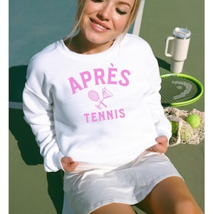 May include: White sweatshirt with pink text that reads "APRÈS TENNIS" and a graphic of a tennis racket and a cocktail glass.