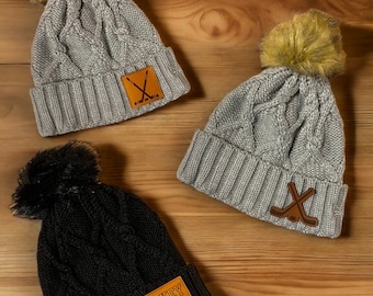 May include: Three grey and black knitted winter hats with pom poms. The hats have a cable knit pattern and feature a leather patch with a hockey stick design. One hat has the text "Hockey Mom" on the patch.