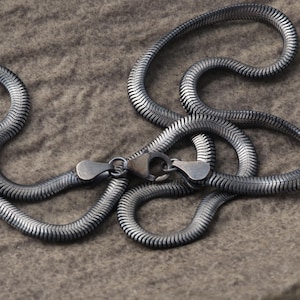 Könnte beinhalten: Eine silberfarbene Schlangenkette mit Karabinerverschluss. Die Kette besteht aus ineinandergreifenden, gerippten Metallgliedern. Die Halskette wird auf einer strukturierten, grauen Oberfläche präsentiert.